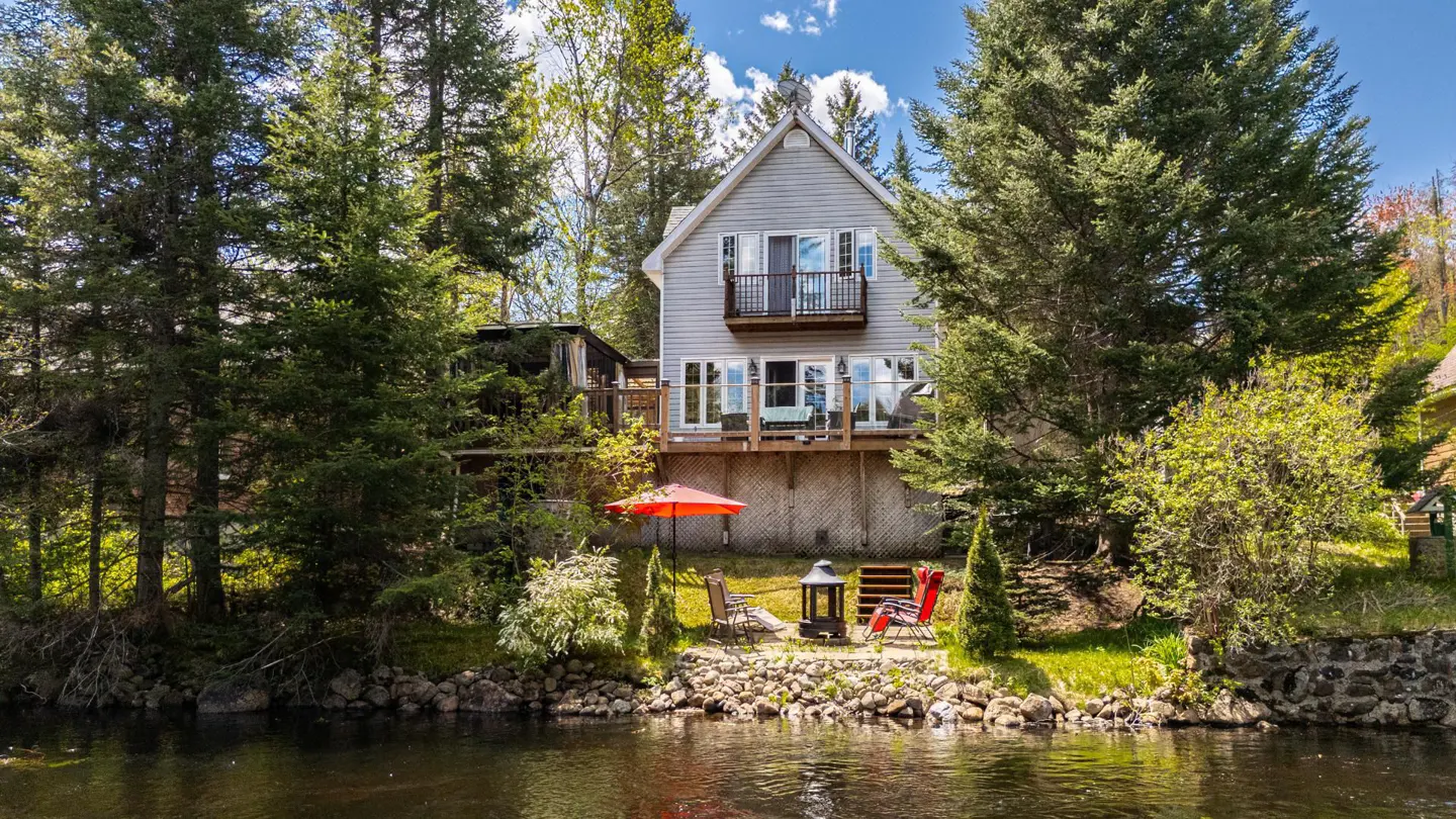 A gray two-story house with a balcony, surrounded by trees, sits on a riverbank with an orange umbrella and chairs.