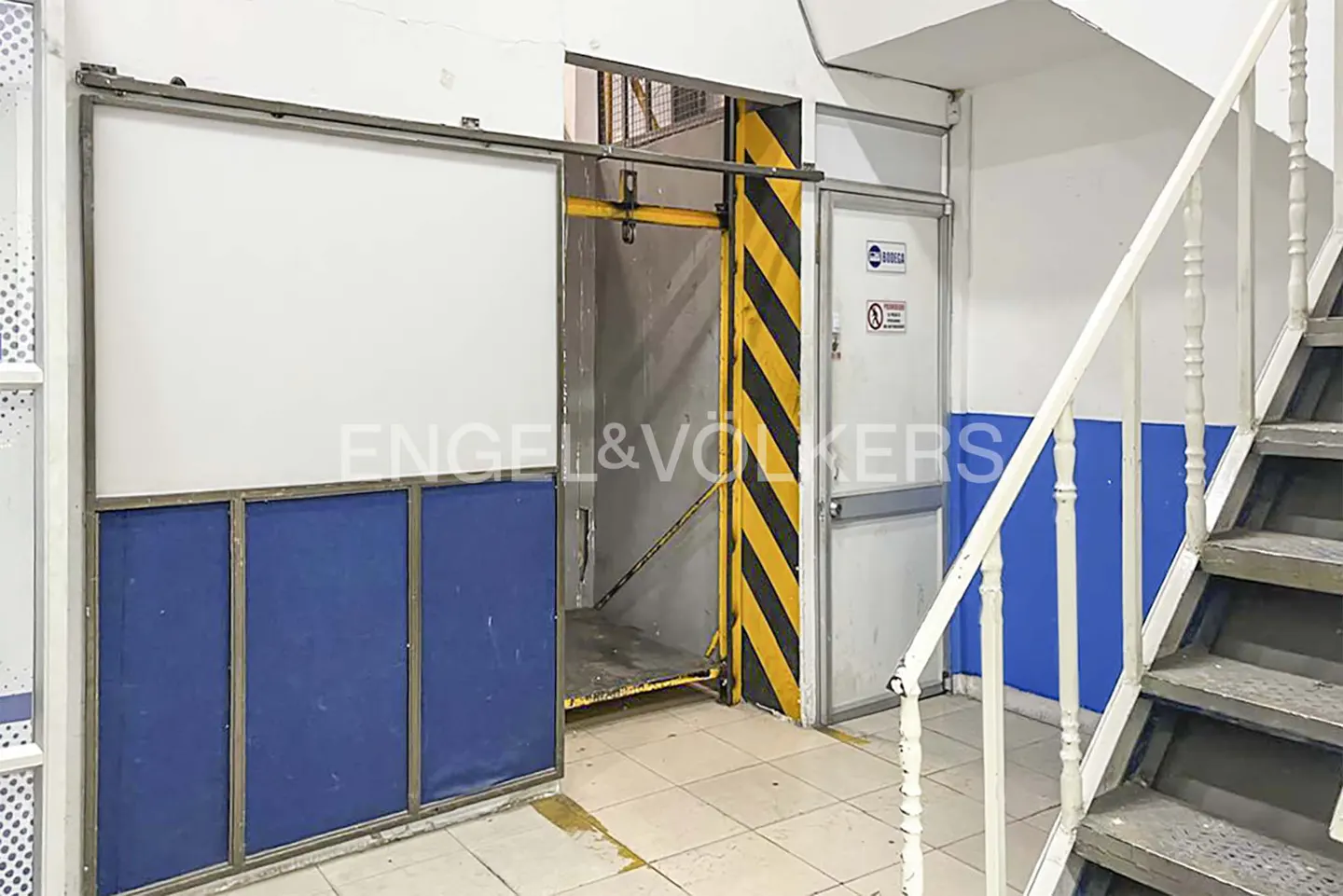 Interior view of a building with a freight elevator, stairs, and a door. The elevator has a blue and white panel and a yellow and black striped frame.