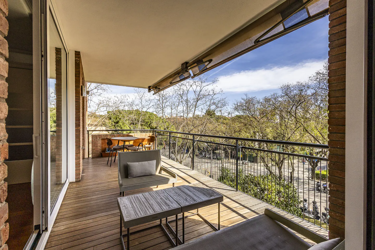 A balcony with a wooden floor, a table, chairs, and a view of trees and a street.