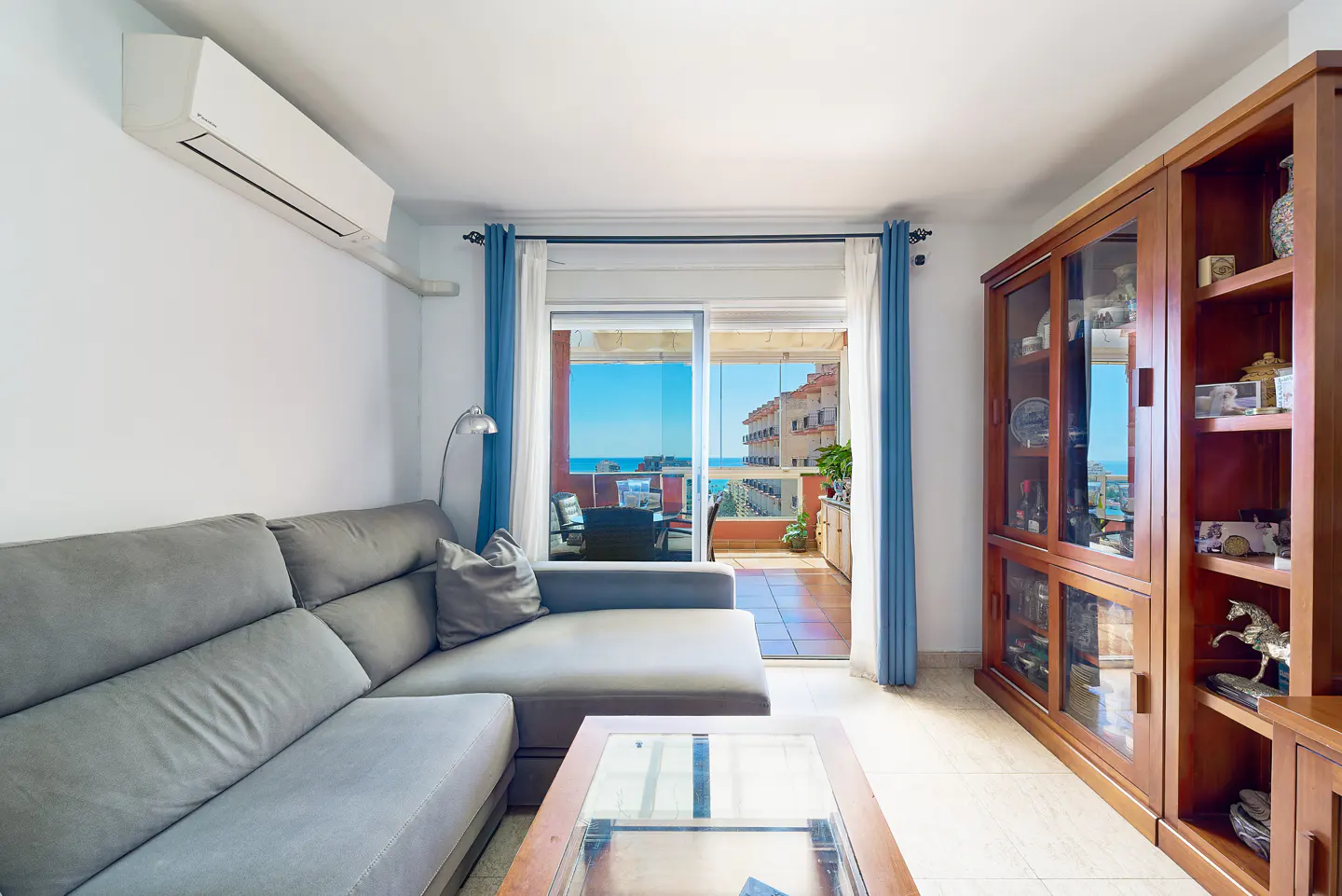 Living room with a gray sectional sofa, glass coffee table, and wooden display cabinet. Balcony with ocean view through sliding glass doors with blue curtains.