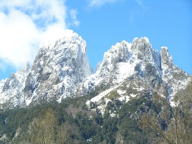 Snow-covered mountain peaks rise against a blue sky with scattered clouds. Green trees cover the lower slopes.