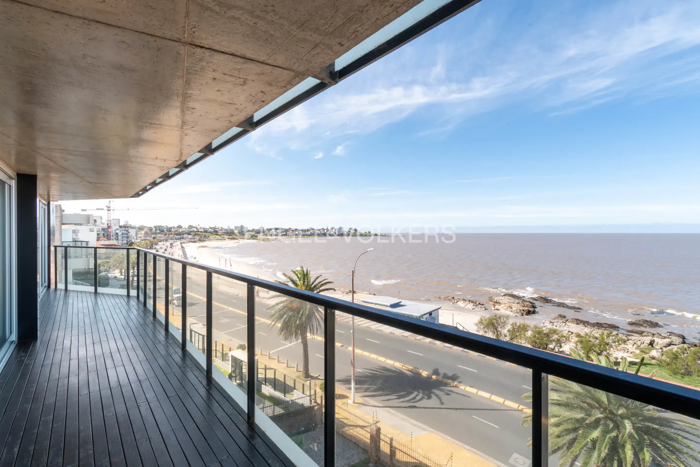 View from a balcony with black wood floors and glass railings, overlooking a beach, road, and ocean under a blue sky.