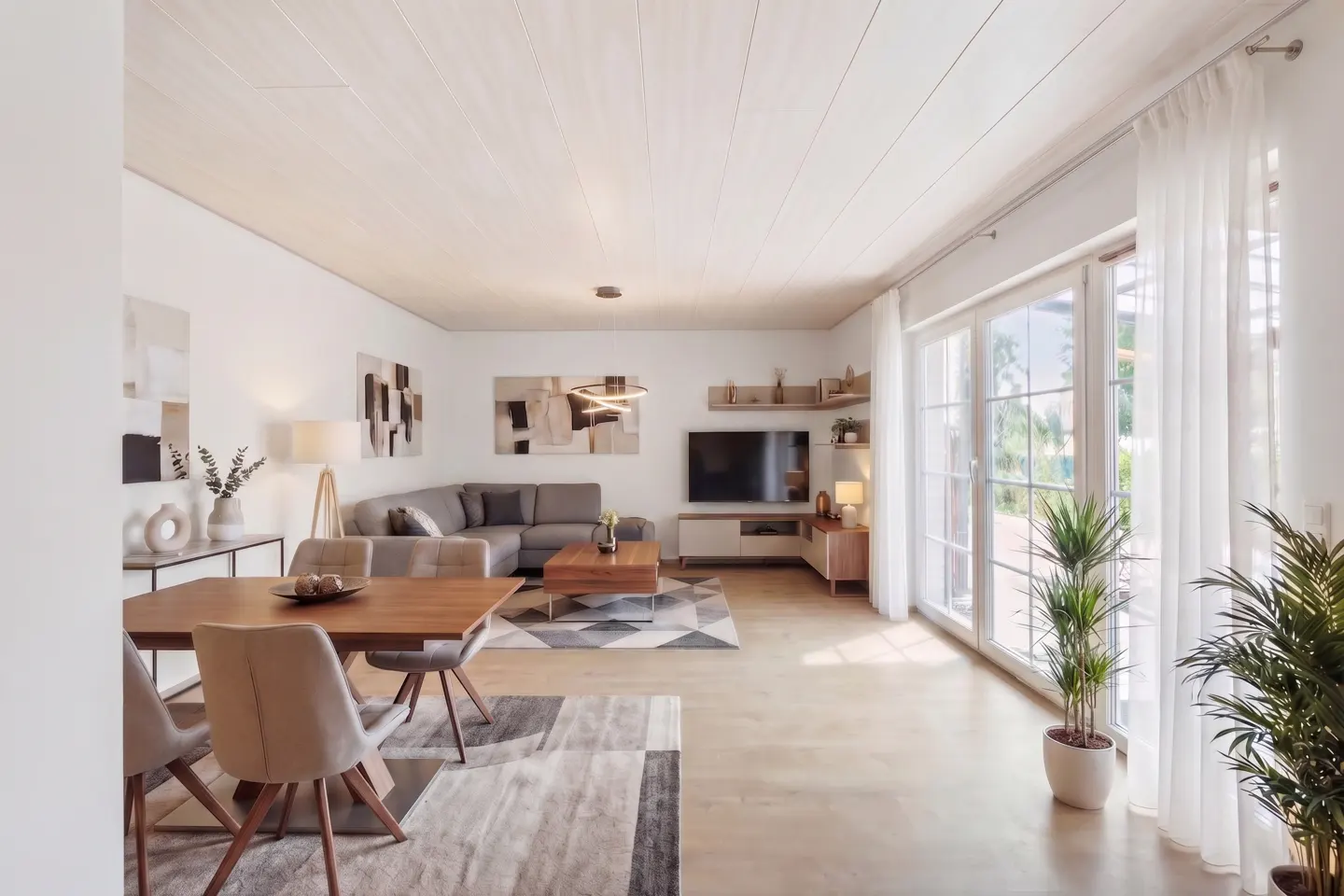 Bright, modern living room with a wood dining table, gray sectional sofa, TV, and large windows with sheer white curtains.