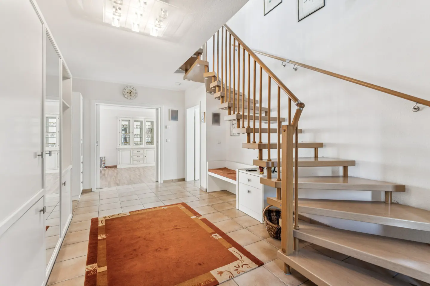 Bright foyer with a wooden staircase, white walls, and beige tile flooring. An orange rug sits in front of white cabinets.