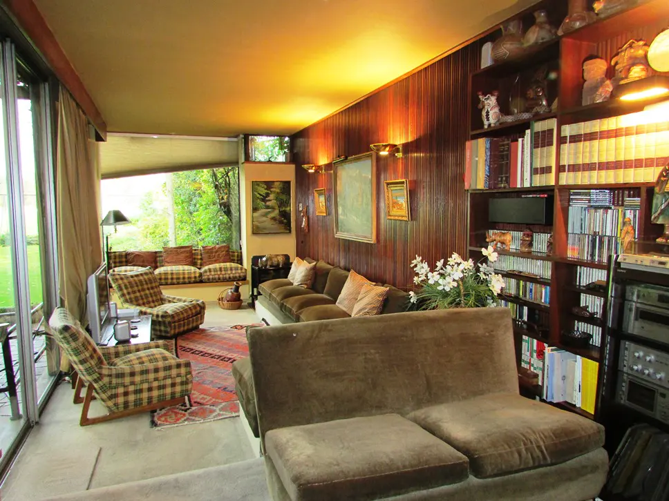 A living room with wood paneling, a bookshelf, and a large window. The furniture is brown and plaid.