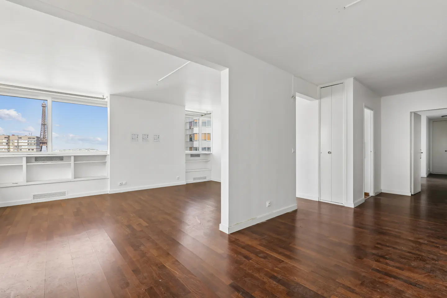 Bright, empty apartment with hardwood floors and white walls. A window shows the Eiffel Tower in the distance.