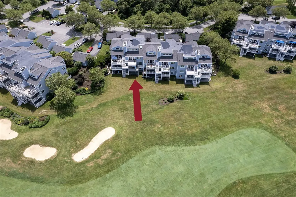 Aerial view of light blue condos with white balconies overlooking a green golf course. A red arrow points to the center condo.