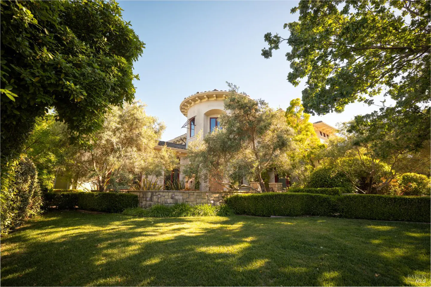 Exterior view of a tan house with a turret, surrounded by trees and a green lawn.