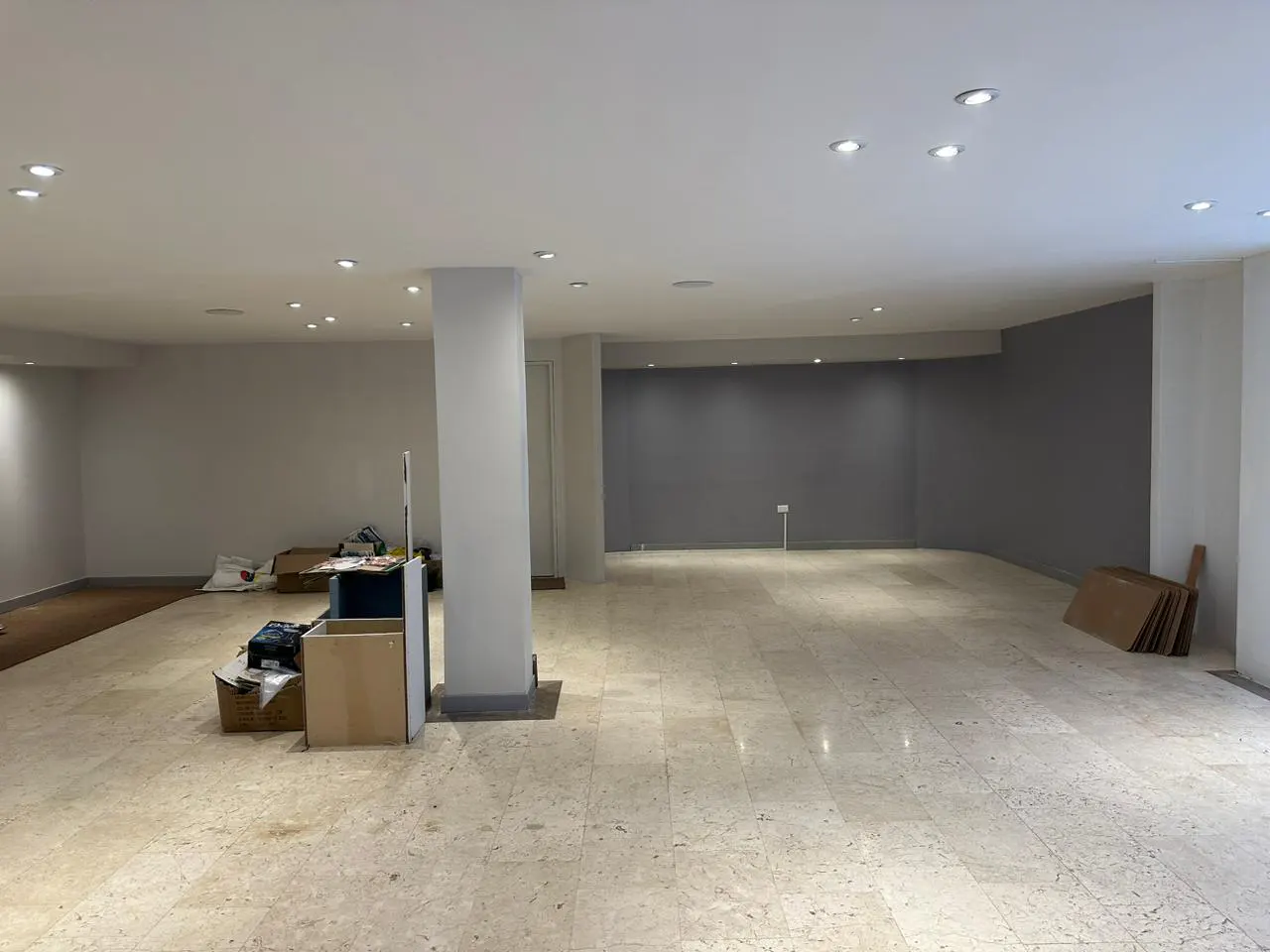 Spacious, empty commercial space with marble floors, white walls, and recessed lighting. Boxes and a support column are visible.