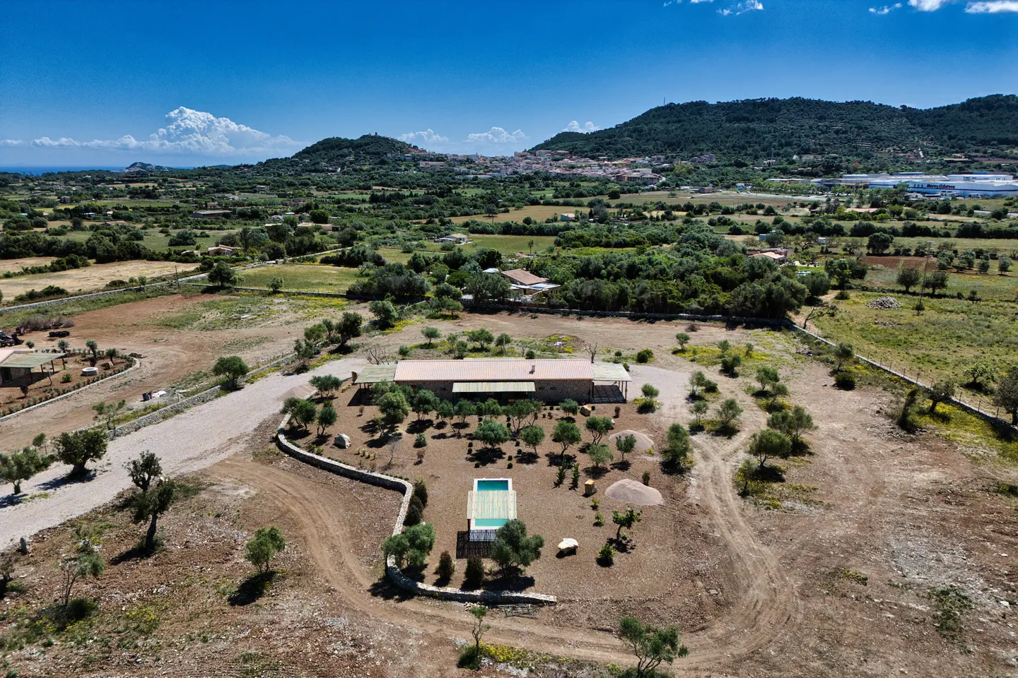 Aerial view of a rural property with a pool, stone house, and olive trees under a blue sky. Rolling hills and a distant town are in the background.