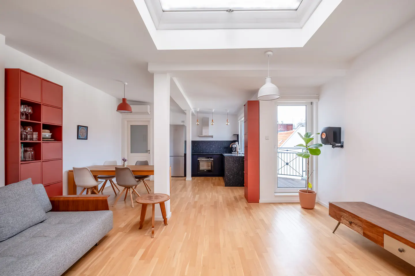 Bright, open-concept apartment with hardwood floors, a gray sofa, and a red bookshelf. A dining table and kitchen are visible in the background.