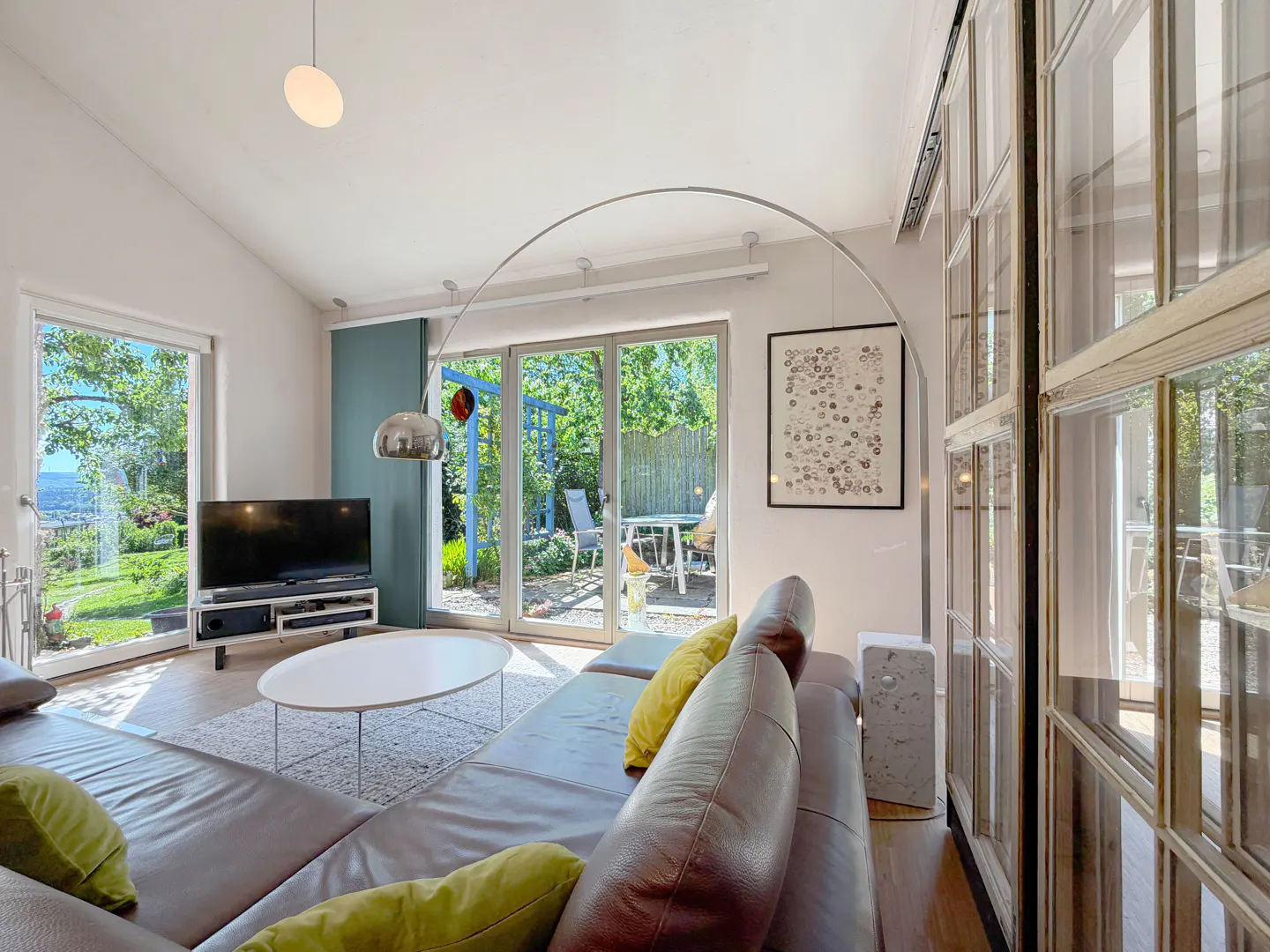 Living room with brown leather sofa, white rug and table, TV, and glass doors to a green garden.
