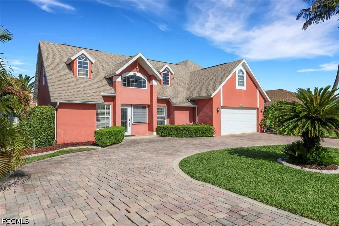 A salmon-colored two-story house with a brick driveway, green lawn, and palm trees under a blue sky.