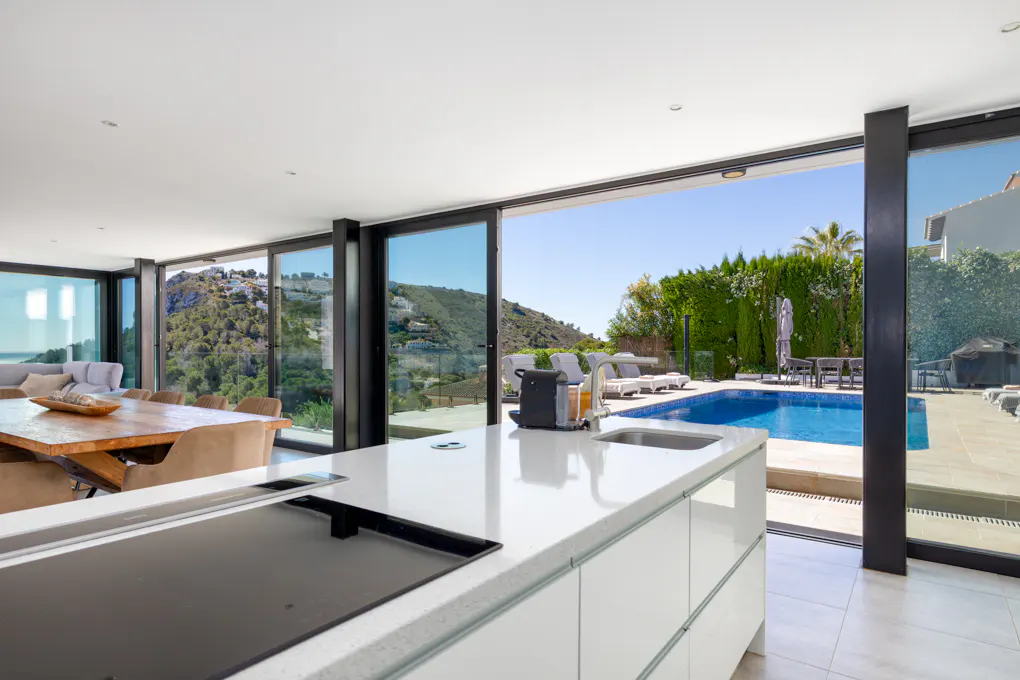 Open-concept kitchen with white cabinets, black cooktop, and sink. Sliding glass doors reveal a pool and mountain view.