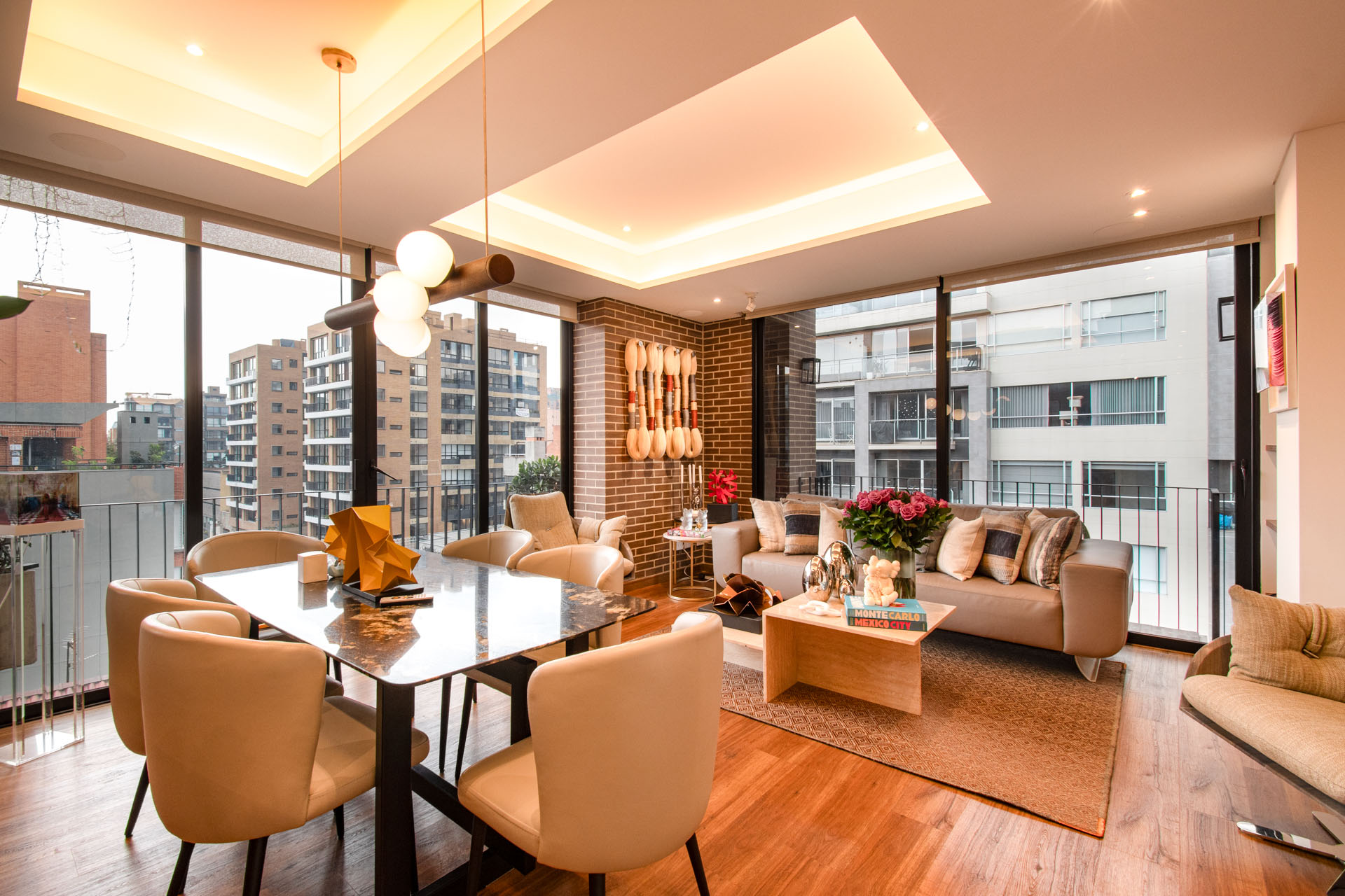 Bright, modern apartment with a dining table, beige chairs, and a sofa. Large windows offer city views. Warm lighting and wood floors create a cozy atmosphere.