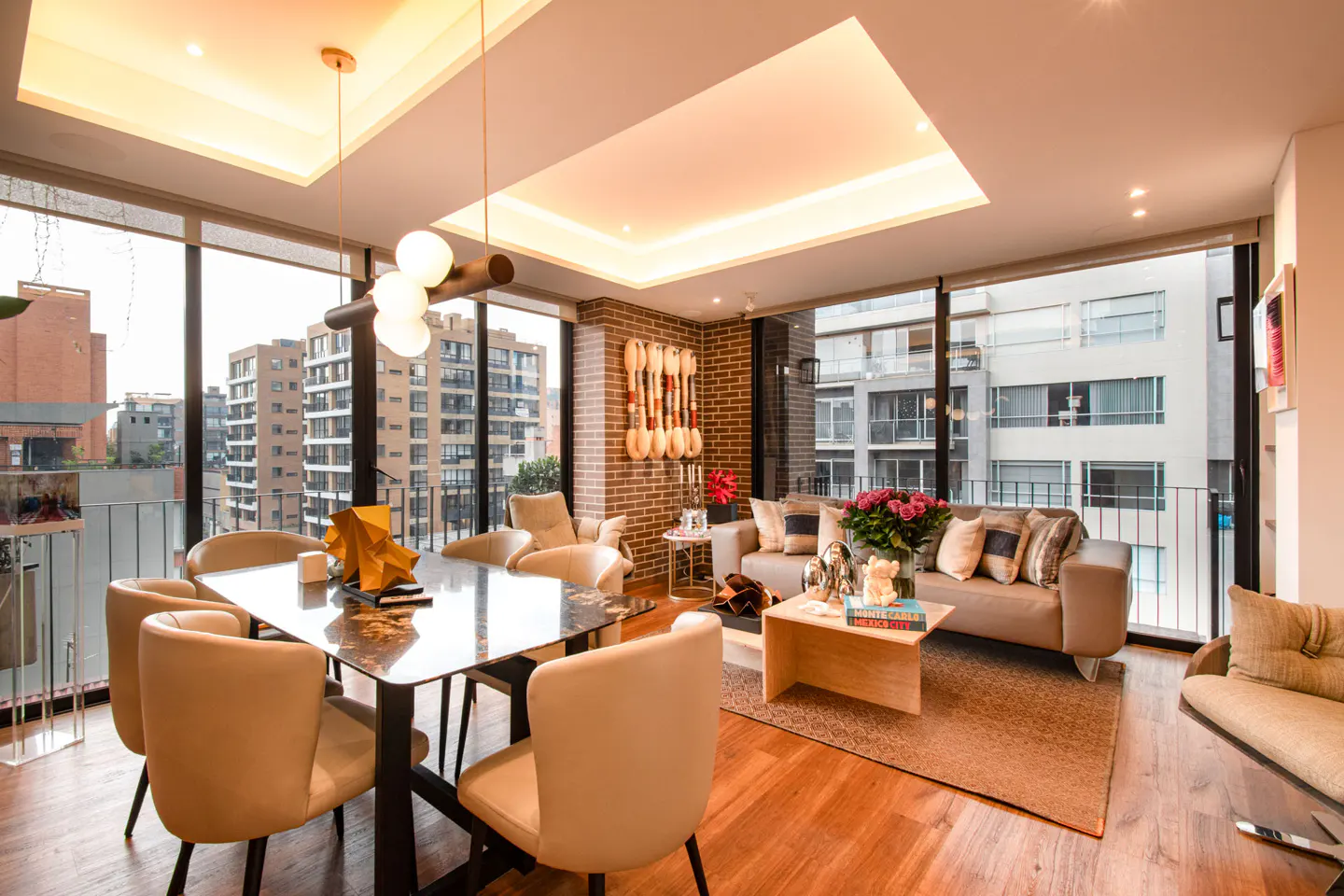 Bright, modern apartment with a dining table, beige chairs, and a sofa. Large windows offer city views. Warm lighting and wood floors create a cozy atmosphere.