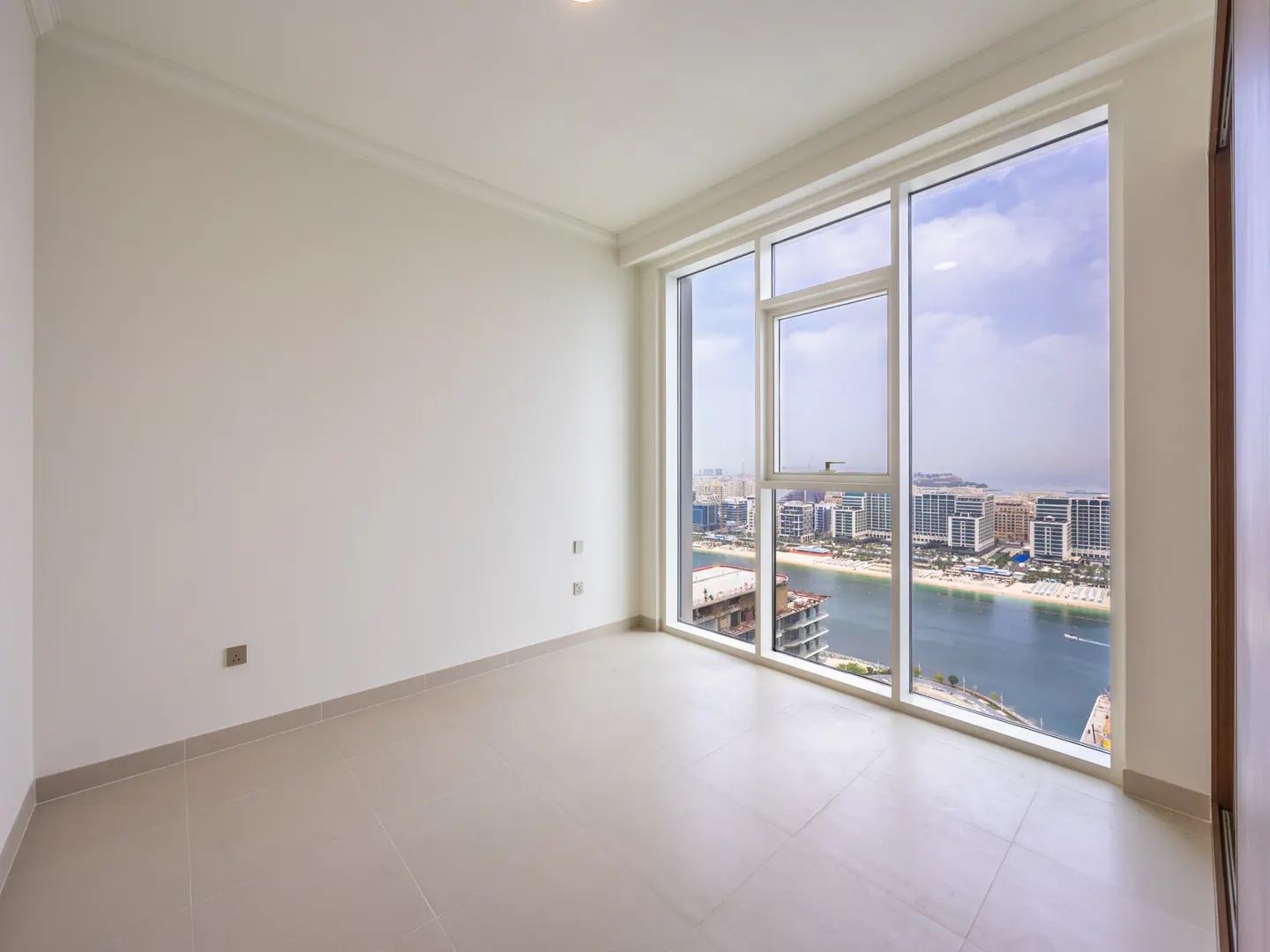 Bright, empty room with white walls and beige tile floor. Large window shows a city skyline, beach, and waterway.