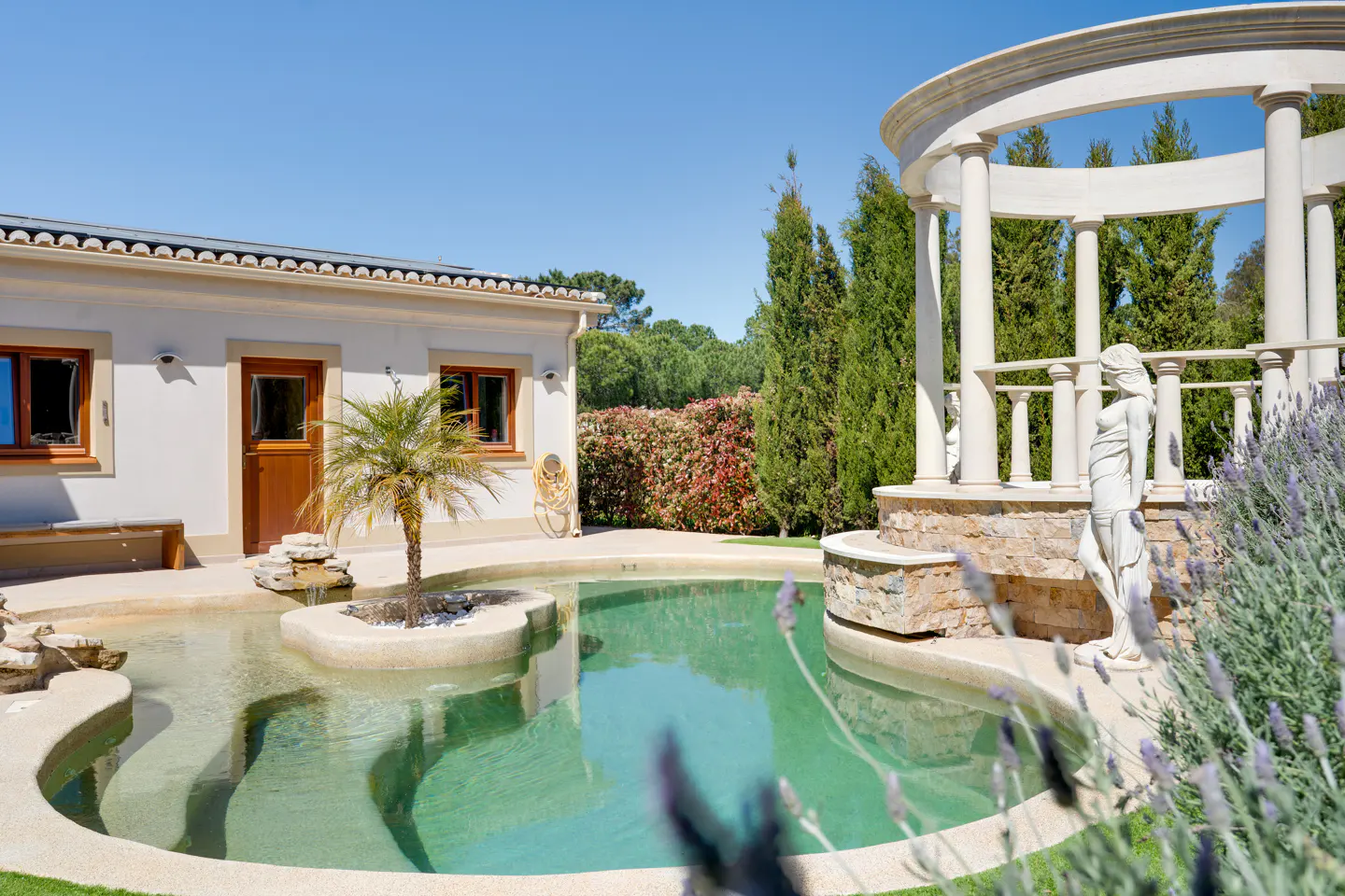 A backyard pool with a small island and palm tree, a white building, and a gazebo with a statue.