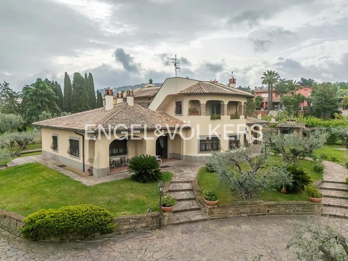 Two-story beige house with a red tile roof, green lawn, and stone walkway on a cloudy day.