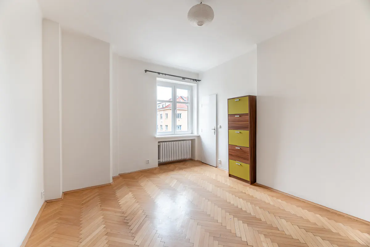 Empty room with white walls, herringbone wood floors, a window with a radiator, and a tall green and wood storage cabinet.