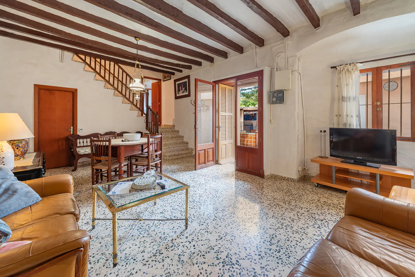 Living room with brown leather sofas, wooden table and chairs, TV, and open door to the outside. Beamed ceiling and tiled floor.