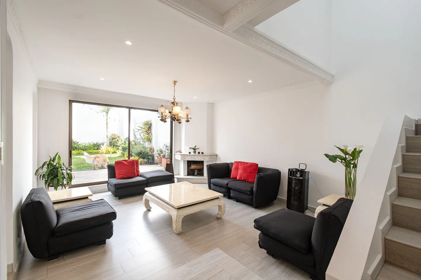 Bright living room with black sofas, red pillows, and a white coffee table. A large window shows a green garden. Stairs are on the right.