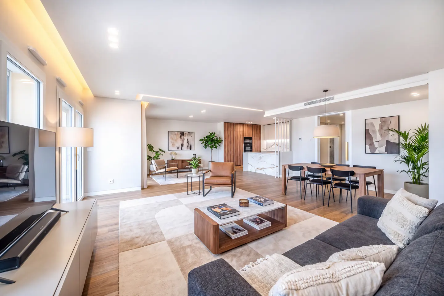 Bright, modern living room with a gray sofa, wood floors, and a dining table. A kitchen is visible in the background.