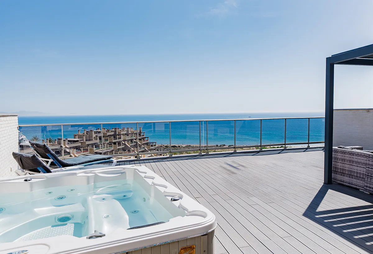 Rooftop patio with a hot tub, lounge chairs, and ocean view. Blue sky and water are visible beyond the glass railing.