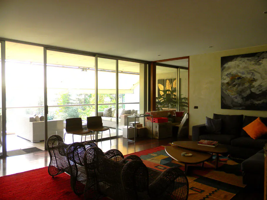 Bright living room with large windows, a gray sofa with orange pillow, and a red rug. A woven chair sits in the foreground.