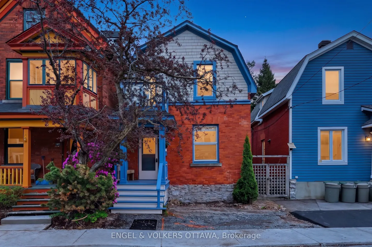 Exterior view of three colorful houses at dusk. The center house is brick with blue trim and a gray shingled upper level.