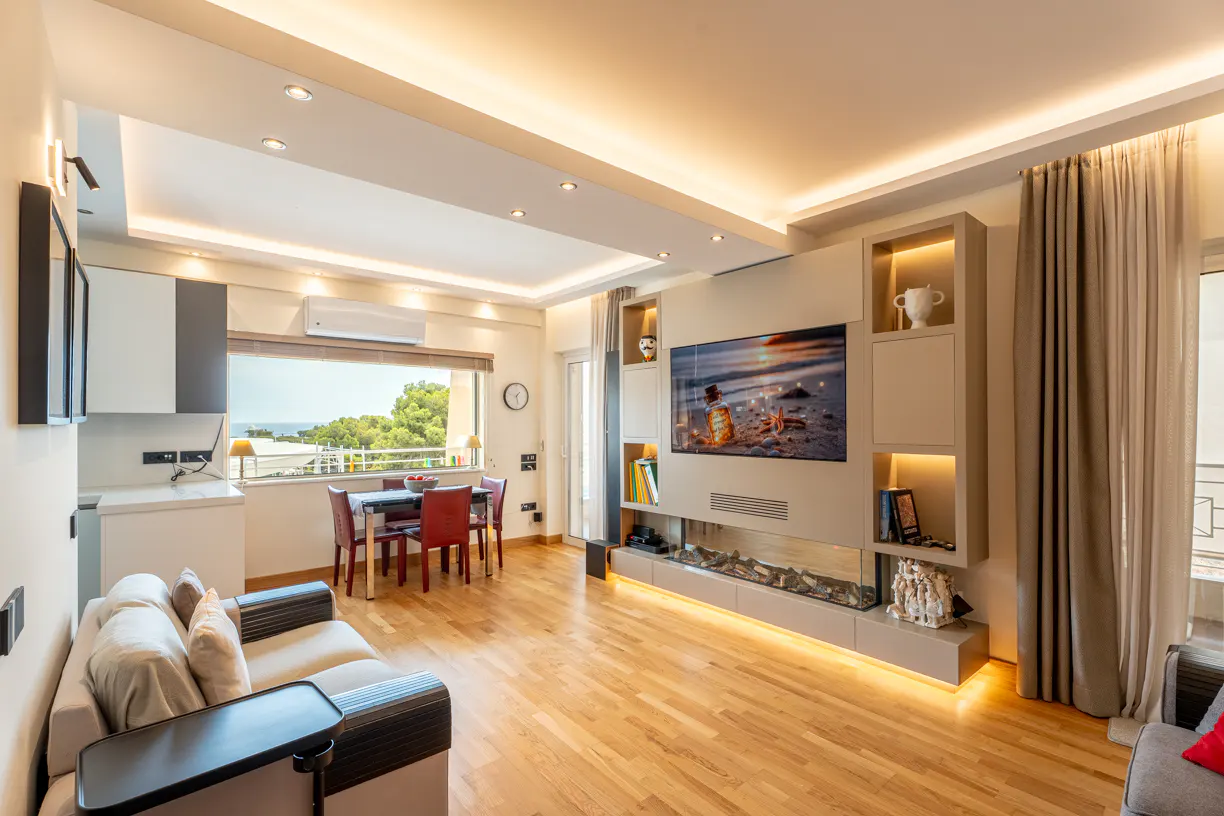 Modern living room with hardwood floors, a wall-mounted TV, built-in shelving, a dining area, and a view of the sea.