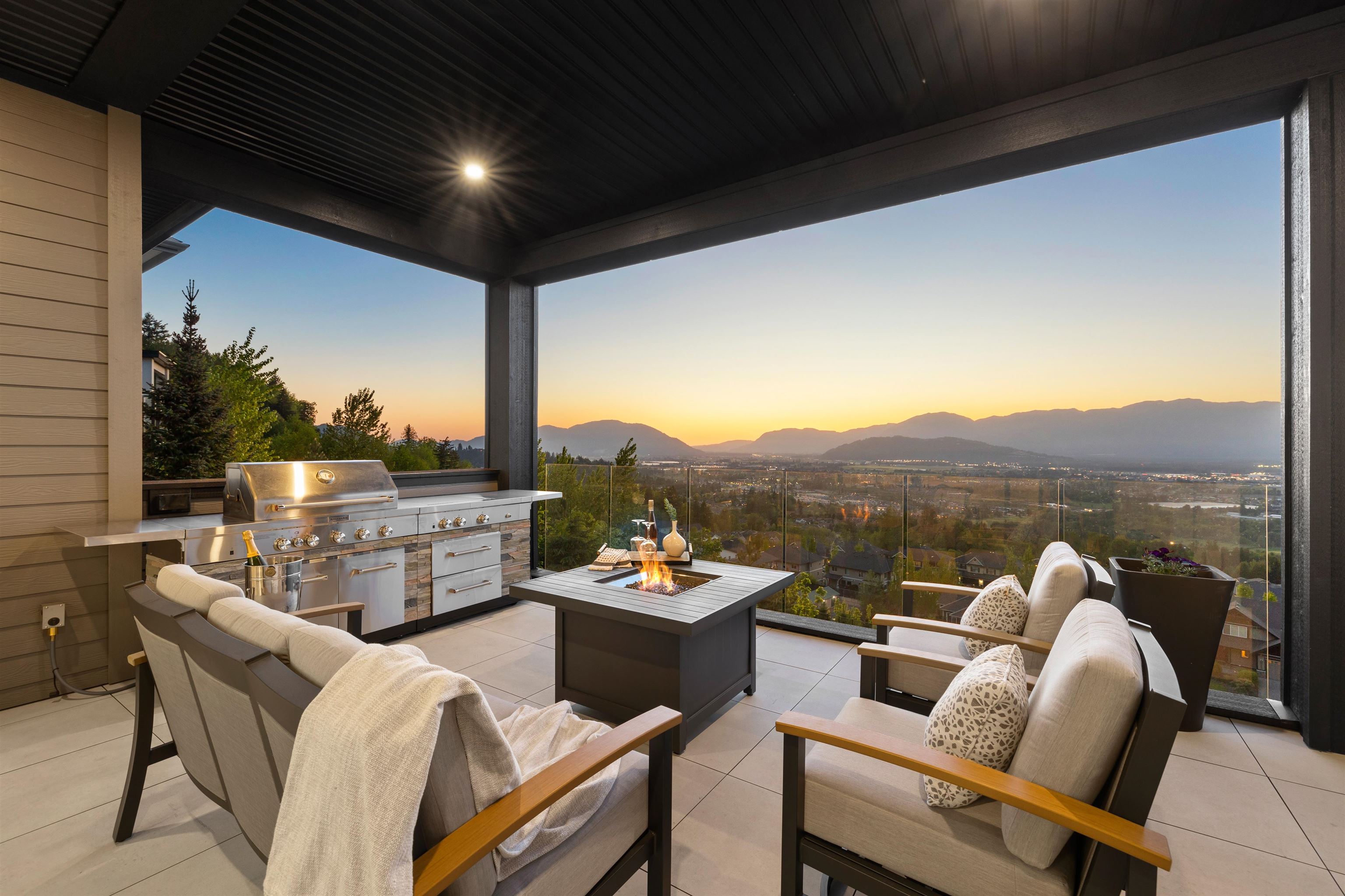 Covered patio with outdoor kitchen, seating, and fire pit overlooking a valley at sunset.