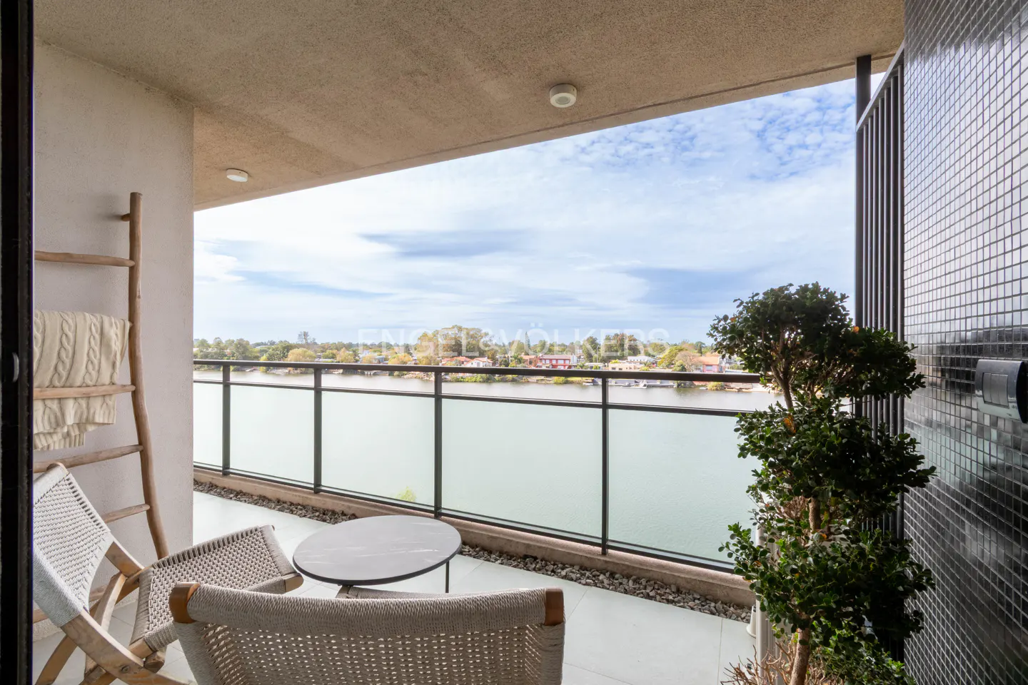 Balcony view with two chairs, a table, and a ladder with a blanket. A river and houses are visible in the background.