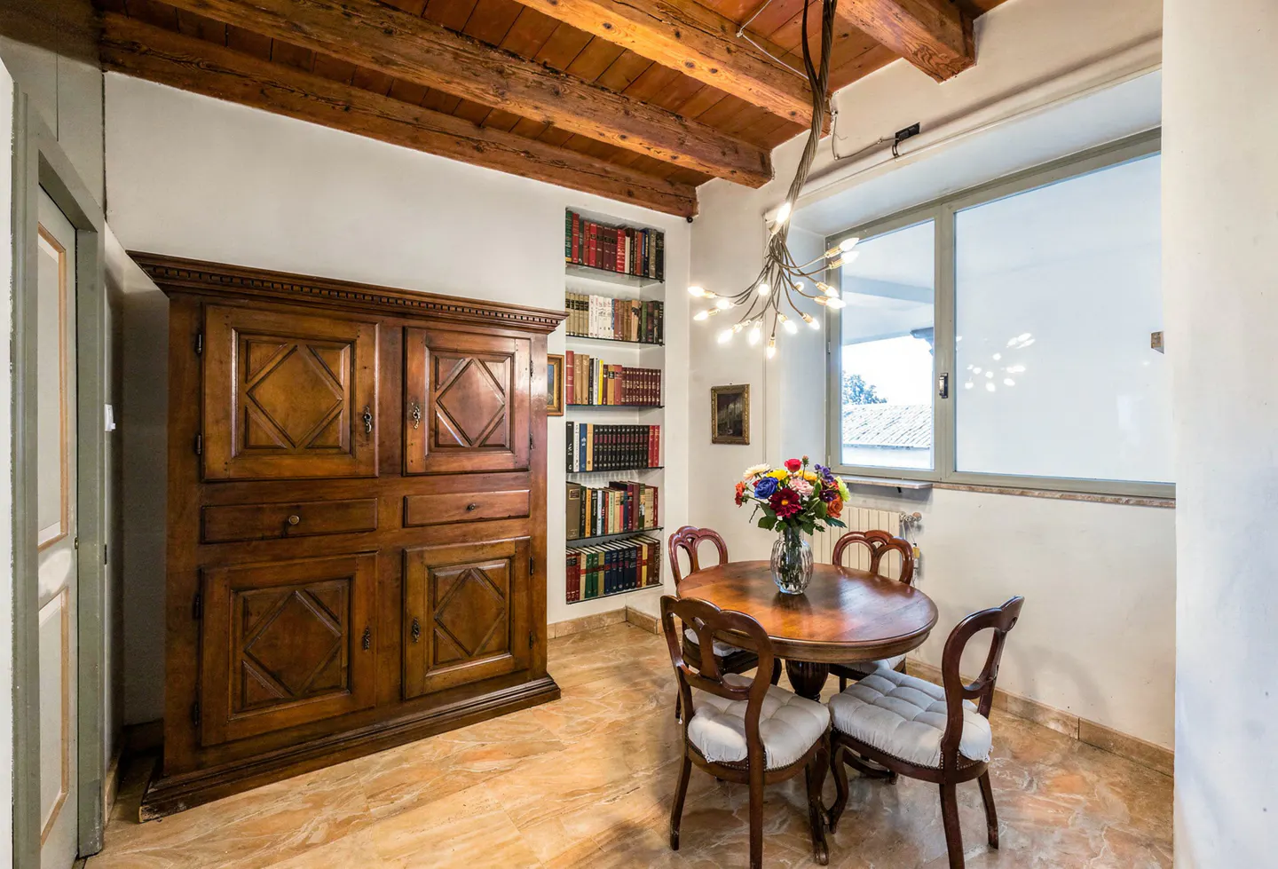 Interior view of a dining area with a wooden table, chairs, and a bouquet of flowers. A large wooden cabinet and a bookshelf are on the left.