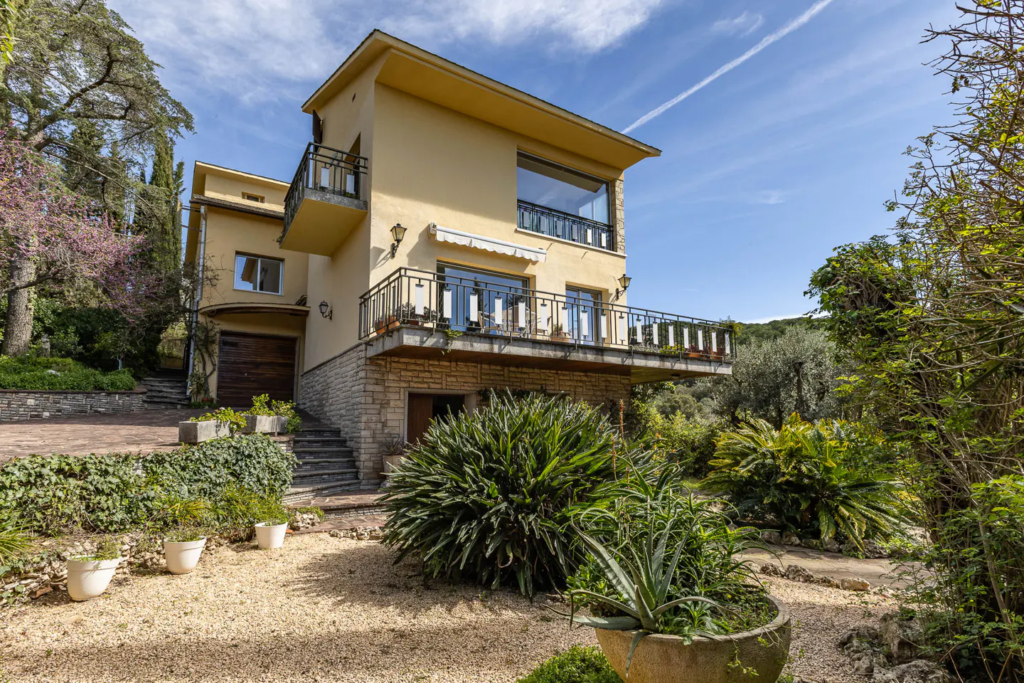 Two-story yellow house with balconies, stone accents, and lush green landscaping under a blue sky.