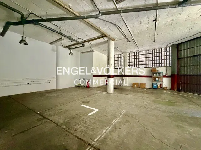 A large, empty commercial space with a concrete floor, white walls, and exposed pipes on the ceiling. A pillar stands in the center.