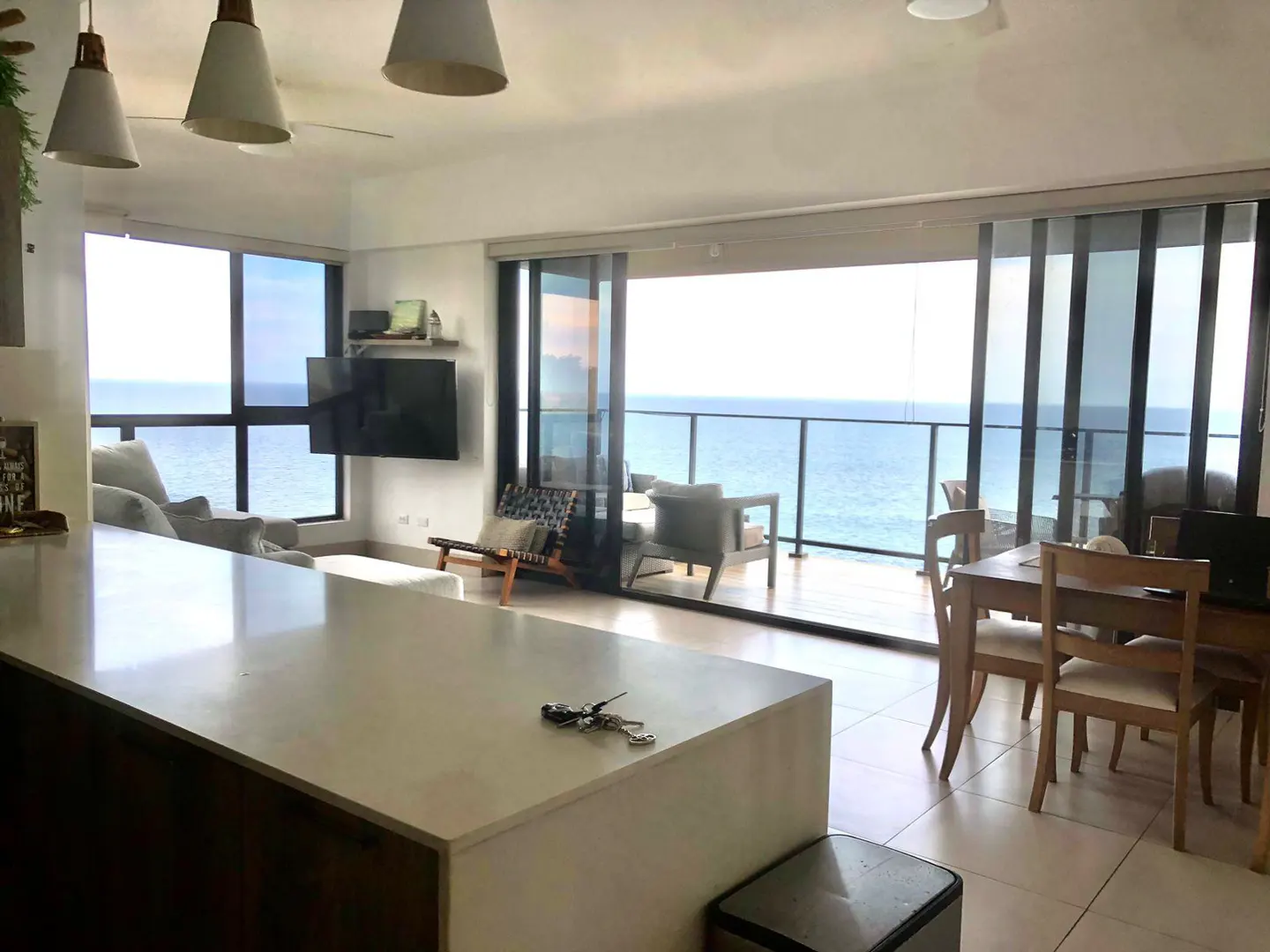 Bright, modern condo interior with ocean view. Kitchen island, dining table, and living area with large windows overlooking the sea.