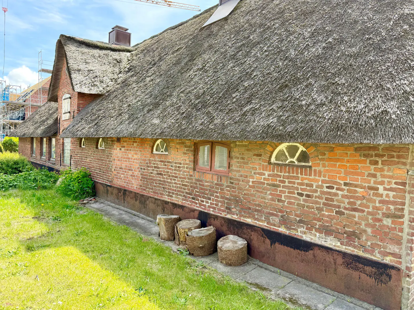 A rustic red brick building with a thatched roof and arched windows, featuring tree stumps on a lawn under a blue sky.