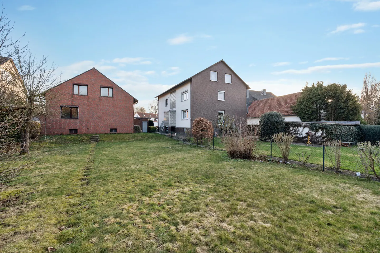 A grassy backyard view featuring a red brick house on the left and a multi-story grey house on the right under a blue sky.