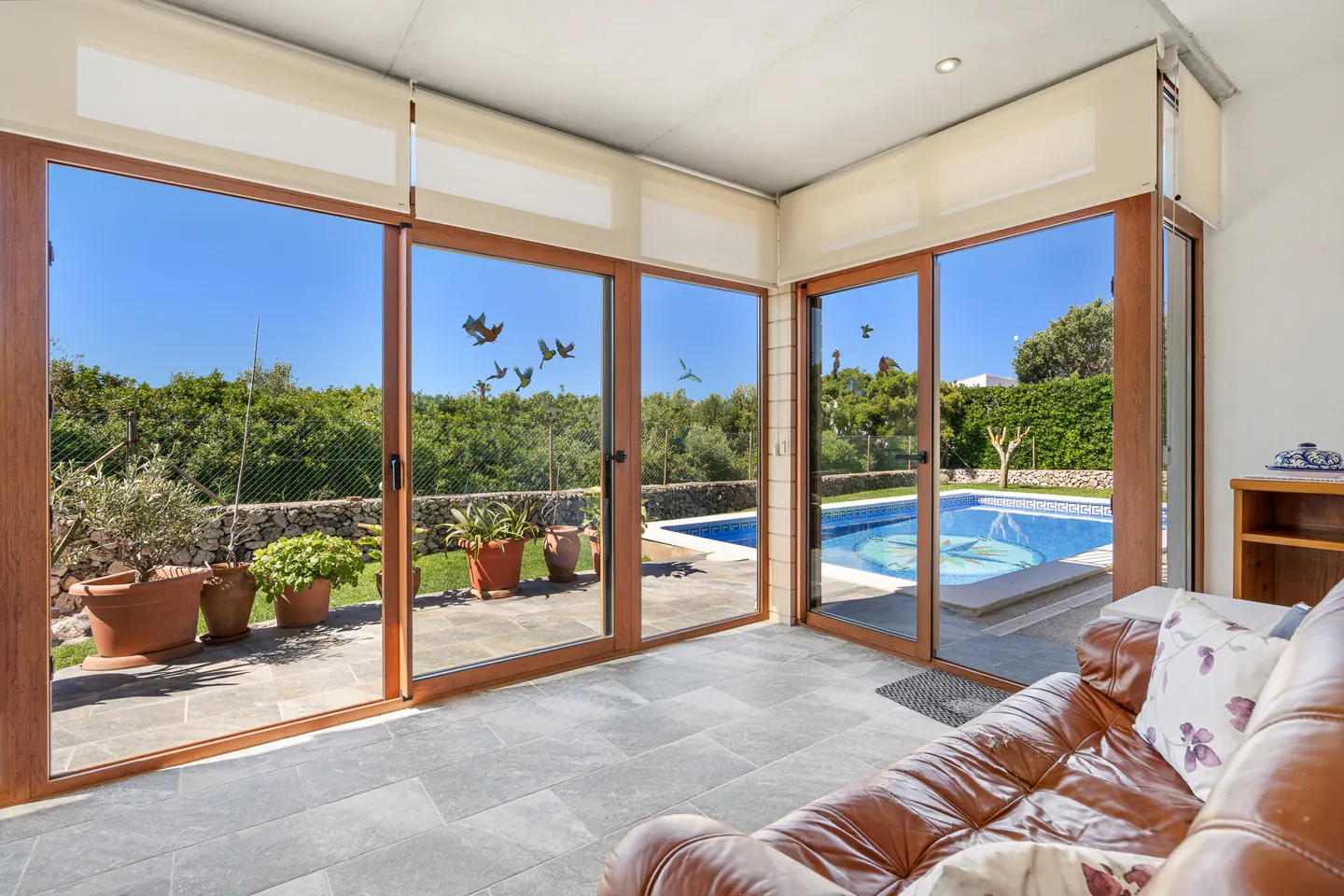 Bright sunroom with brown framed sliding glass doors opening to a pool and garden. A brown leather couch sits on gray tile.