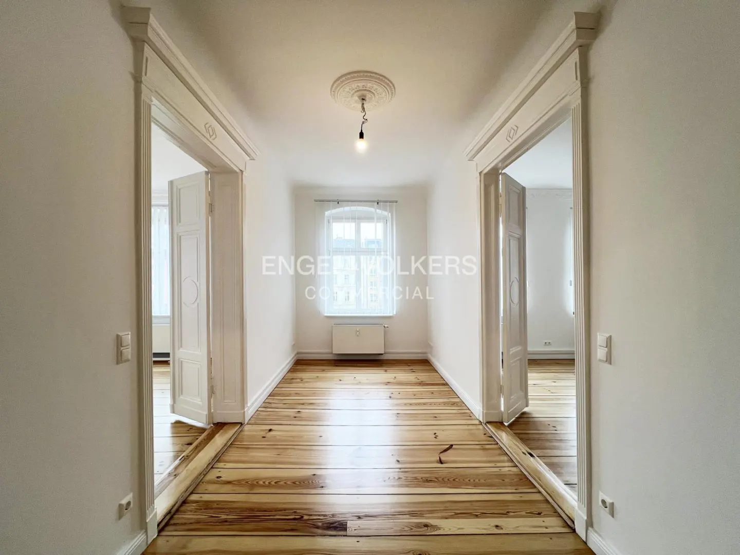 Hallway with light wood floors, white walls, and two open doorways. A window is at the end of the hall with a light fixture on the ceiling.