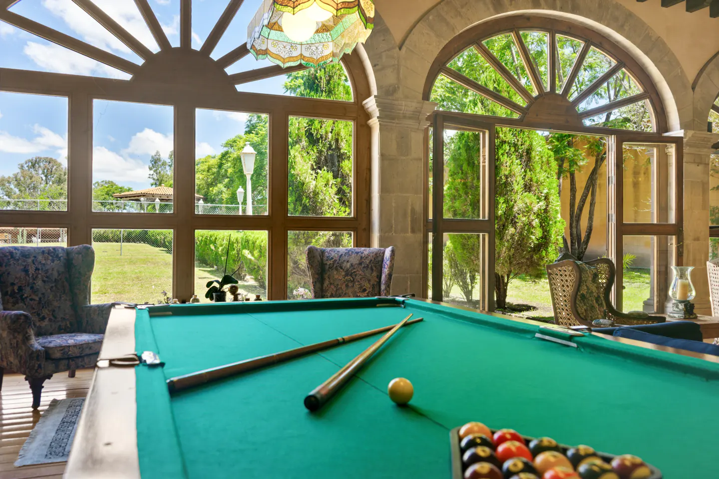 Indoor game room with a green pool table, cues, and balls. Large windows overlook a green lawn and trees.
