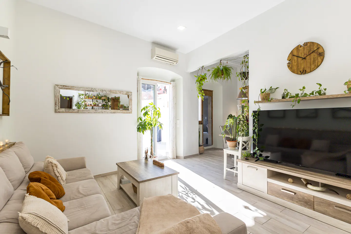 Bright living room with a beige sofa, wooden coffee table, TV, and many plants. Sunlight streams in through an open door.