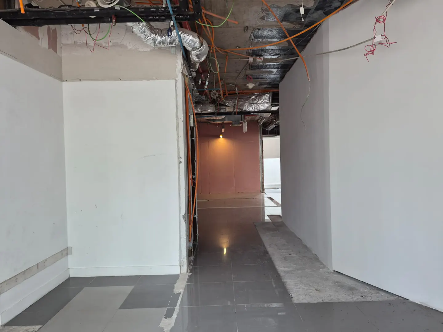 Interior view of a commercial space under construction, featuring white walls, gray tile flooring, and exposed ceiling with ductwork and wiring.