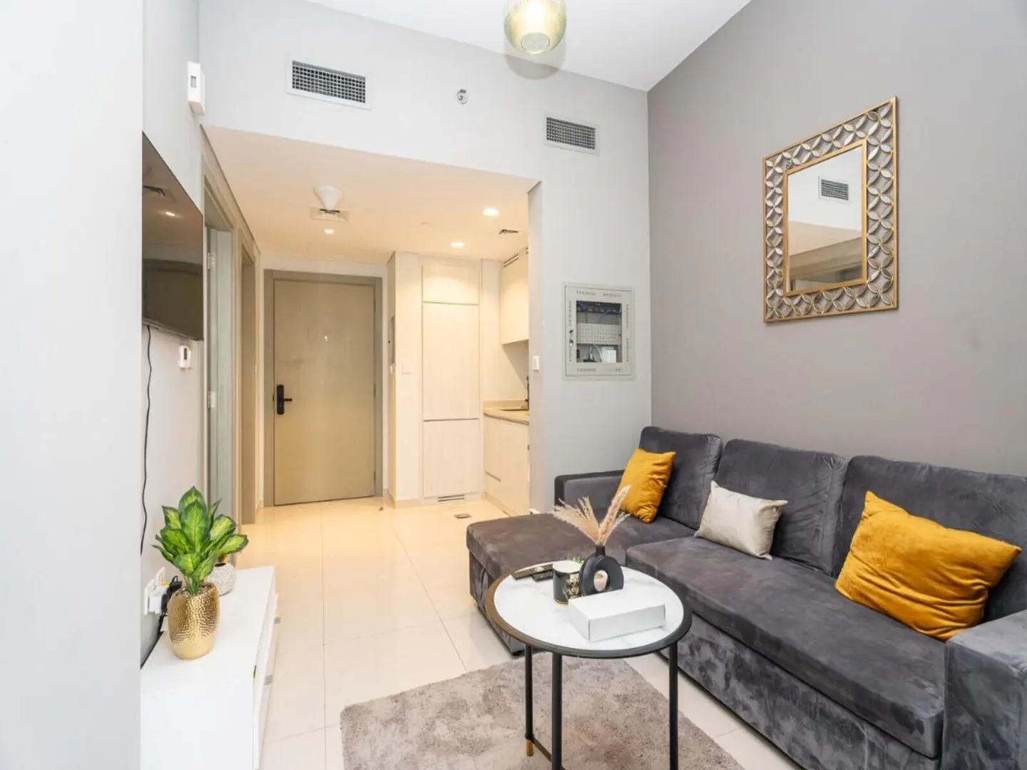 Living room with gray sofa, yellow pillows, and round table. A mirror hangs on the gray wall. A doorway leads to a white kitchen.