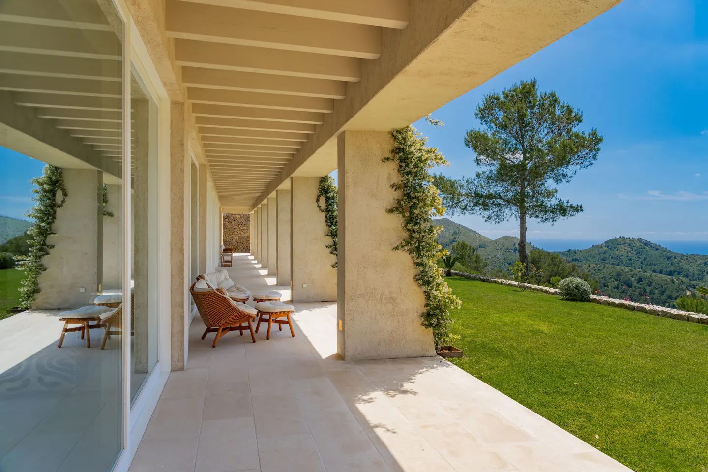 Covered patio with wicker chairs and a view of green hills and blue sky. Vines climb the beige columns.