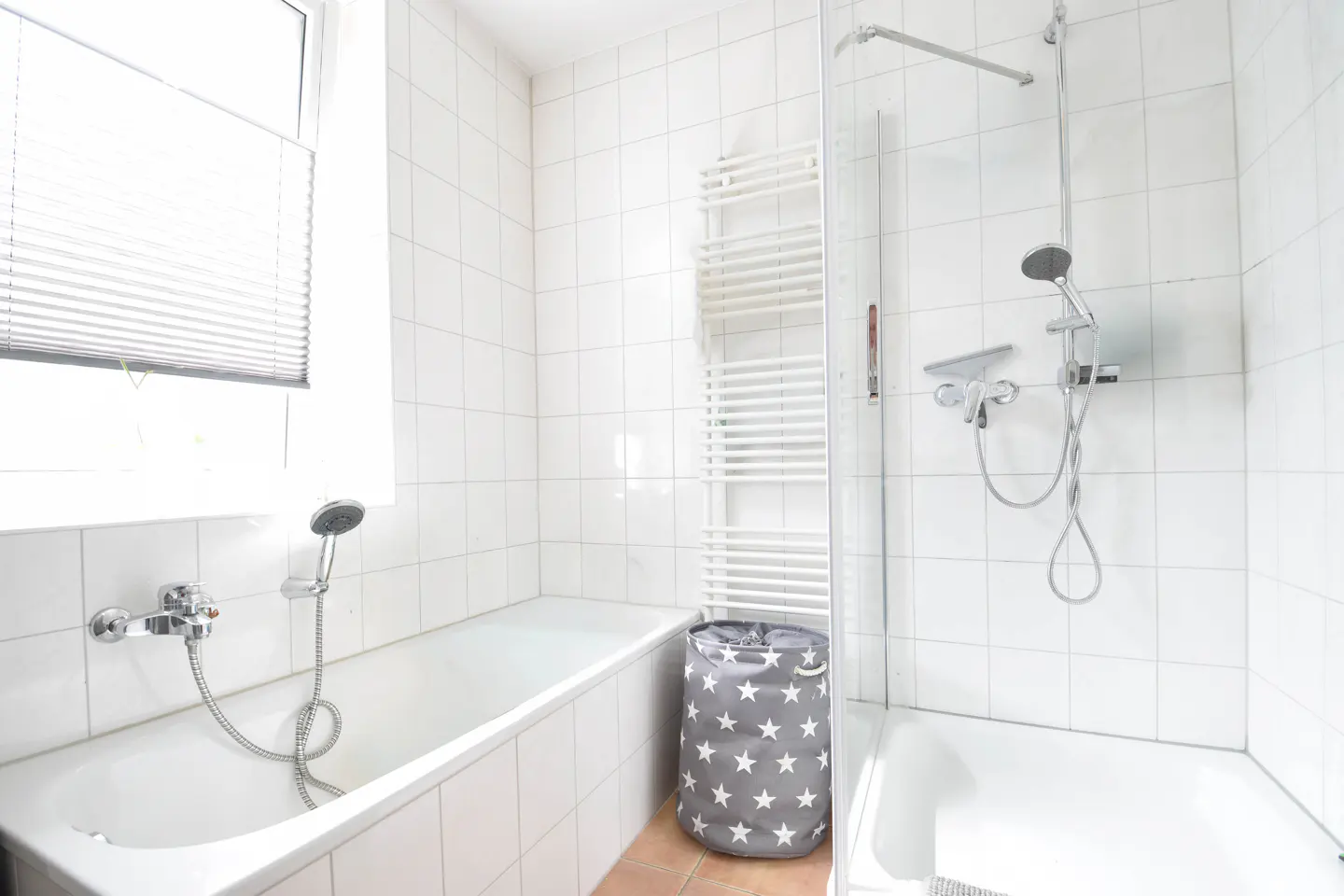 Bright bathroom with white tiled walls, a bathtub with shower, and a gray laundry basket with white stars.