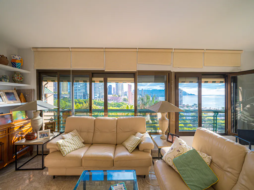 Living room with beige sofas, pillows, lamps, and a glass table. Large windows offer a city and ocean view. Shelves display decorative items.