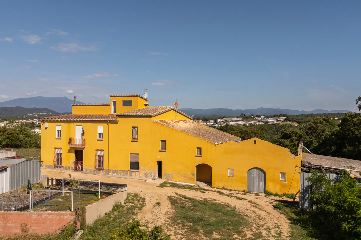 A yellow, two-story farmhouse with a tile roof sits on a dirt road under a clear blue sky.