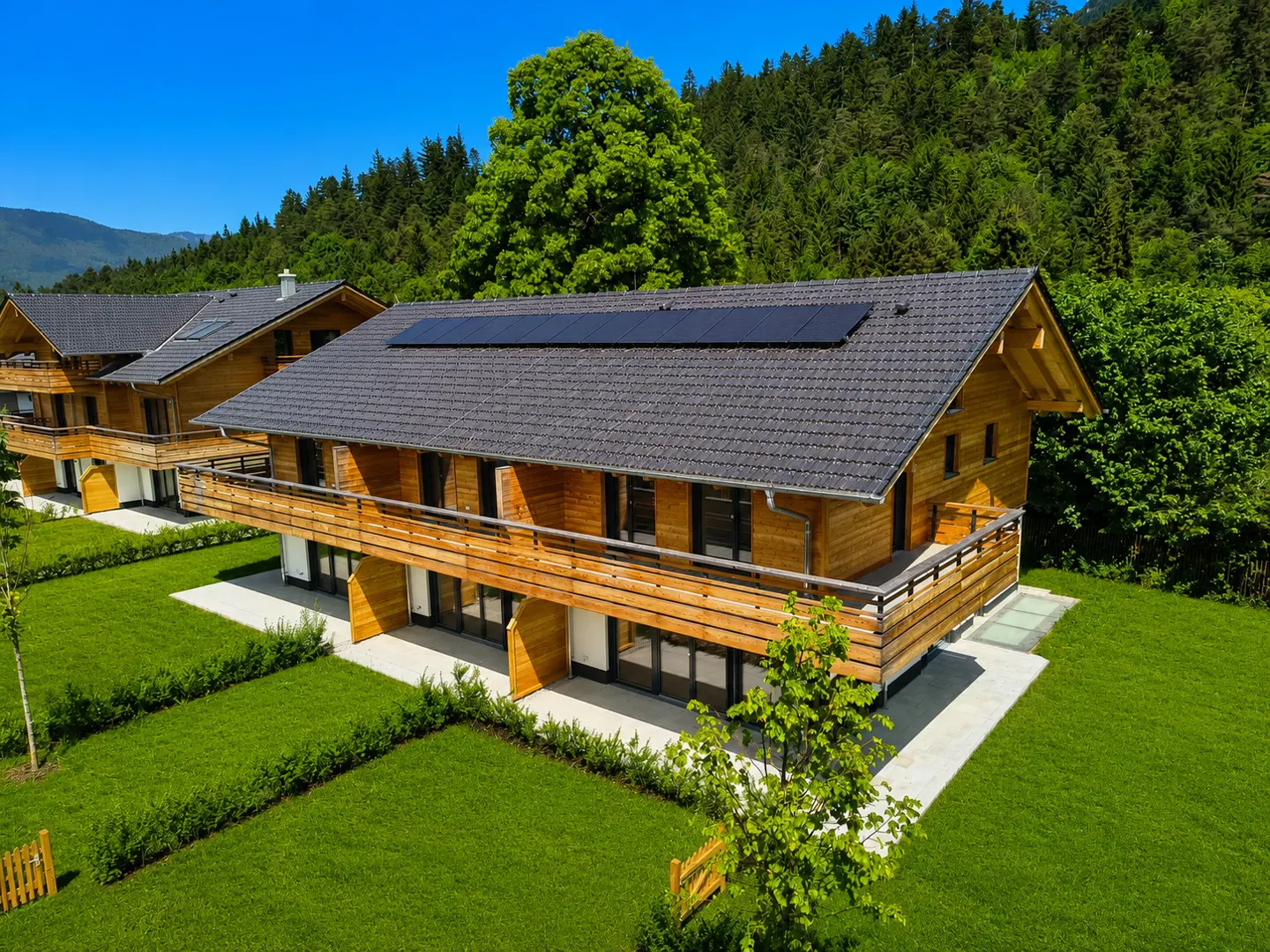 Aerial view of a modern wooden chalet with a dark tiled roof, solar panels, and a balcony, set against a lush forest.